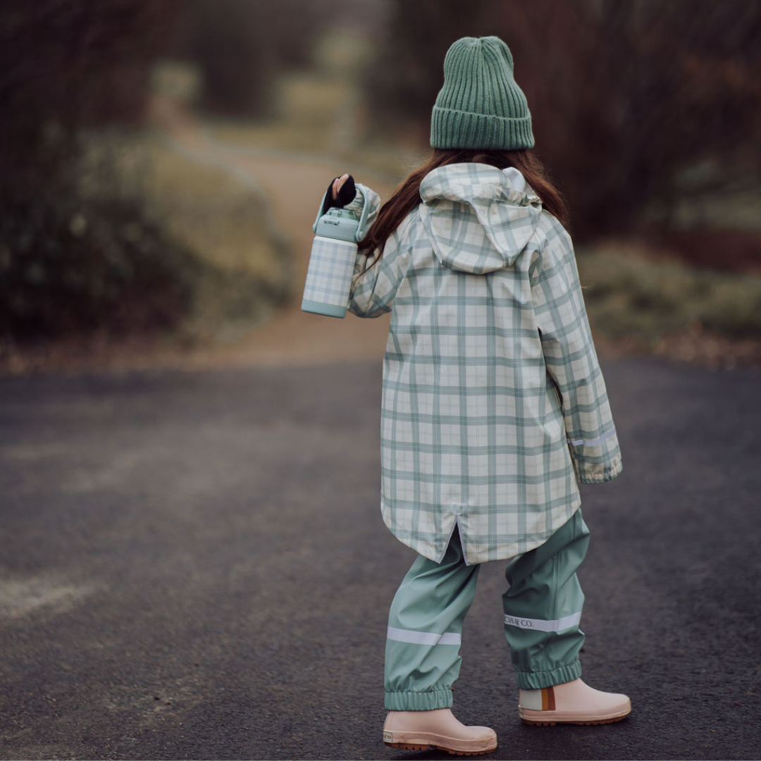Person wearing a green plaid coat and hat, walking on a path with a blurred natural background