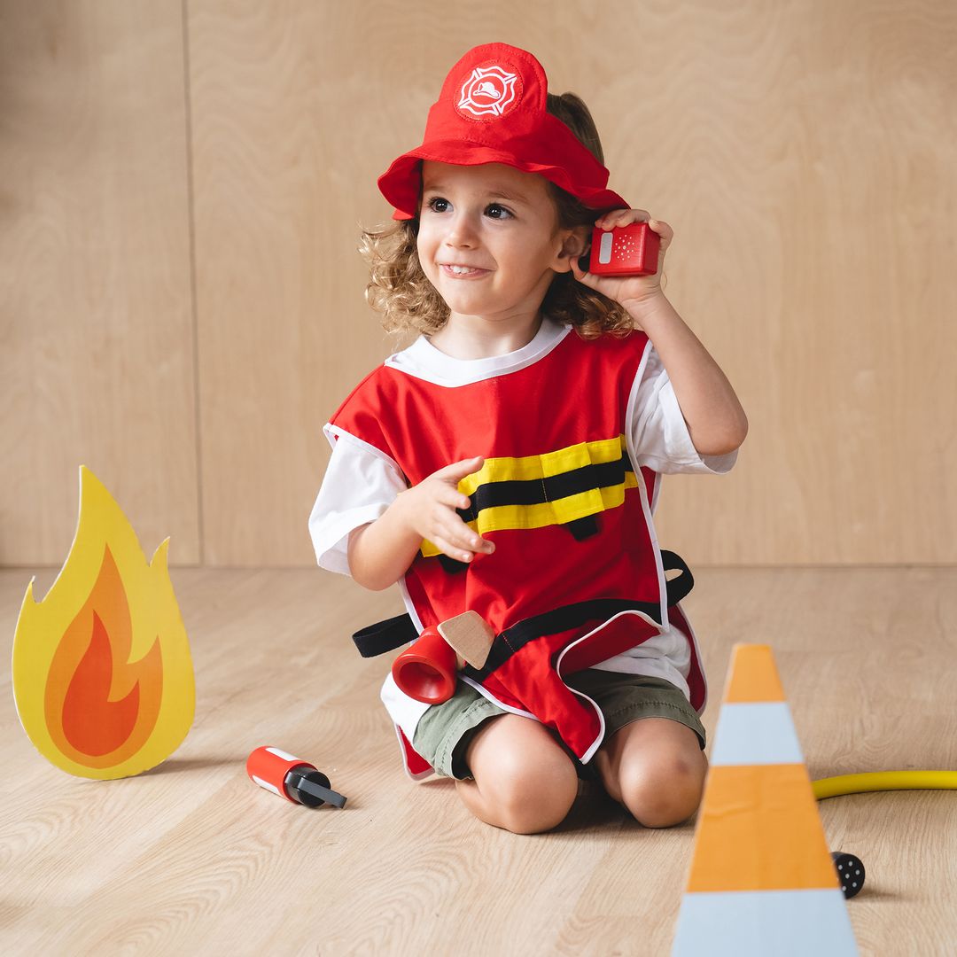 Child in a fireman costume with a toy fire truck, fire extinguisher, and traffic cone on a wooden floor.