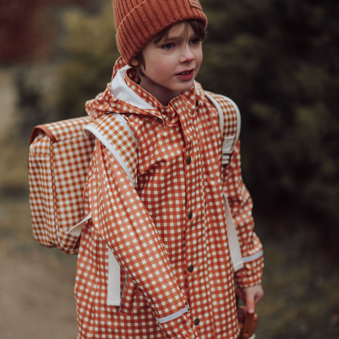 Child wearing an orange checkered raincoat and hat with a backpack outdoors.