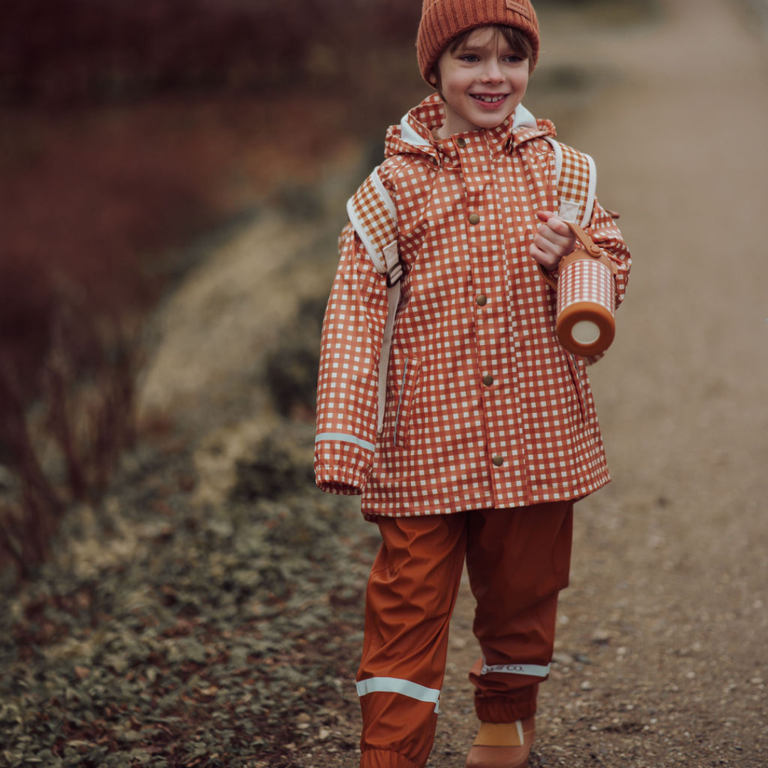 Child in a red checkered raincoat and orange pants holding a roll of paper in a natural setting.
