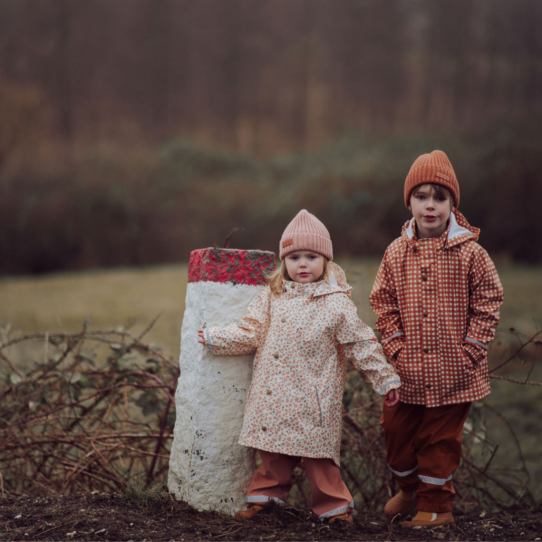 Two children in autumn clothing standing near a stone structure with a red top in a field.