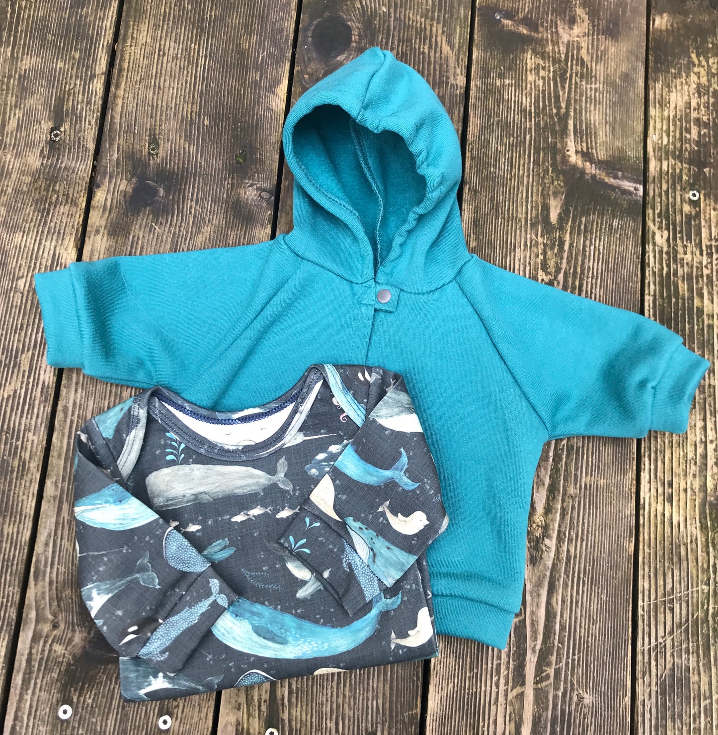 Teal hoodie and whale-patterned onesie on a wooden surface