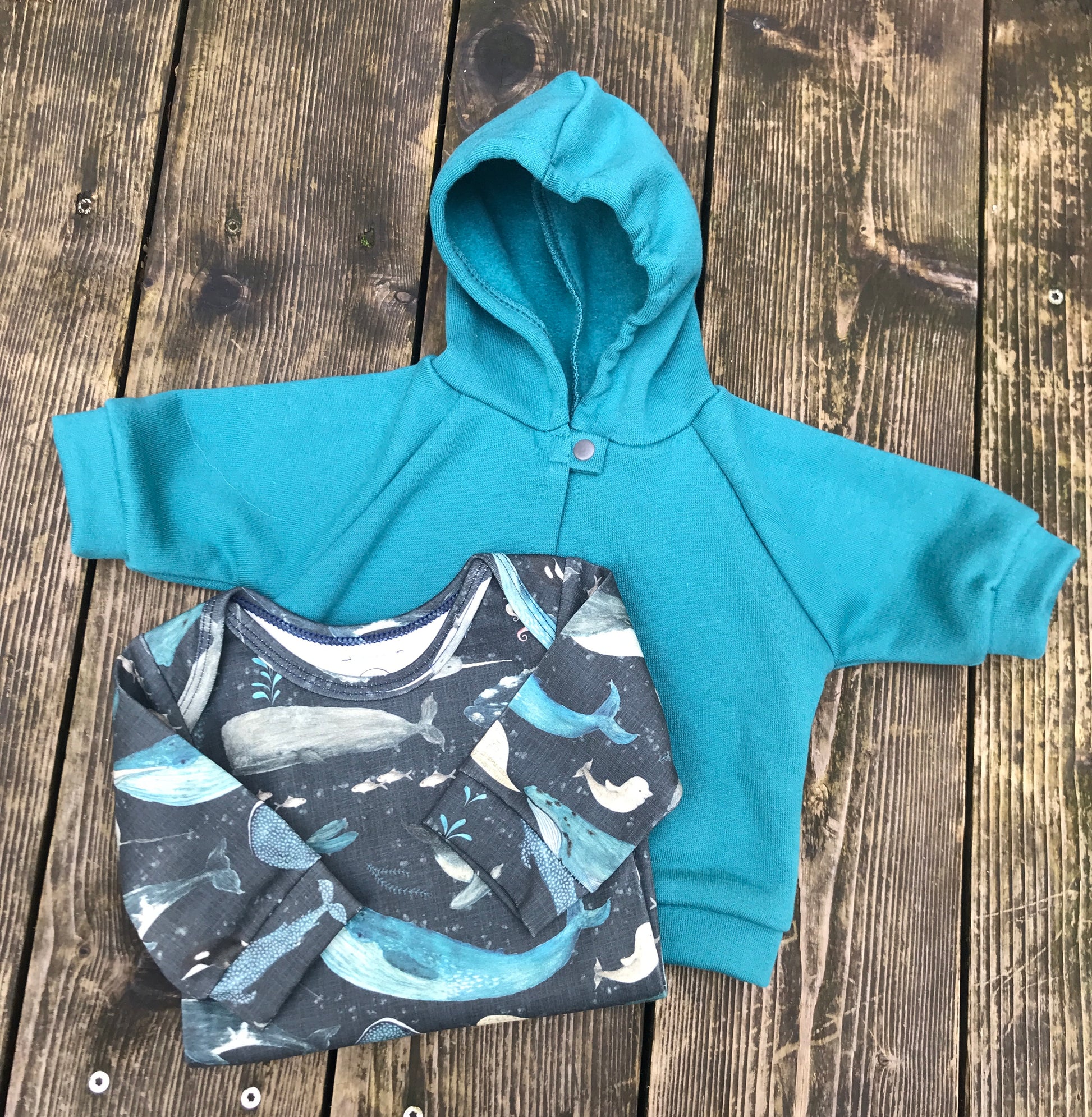 Teal hoodie and whale-patterned onesie on a wooden surface
