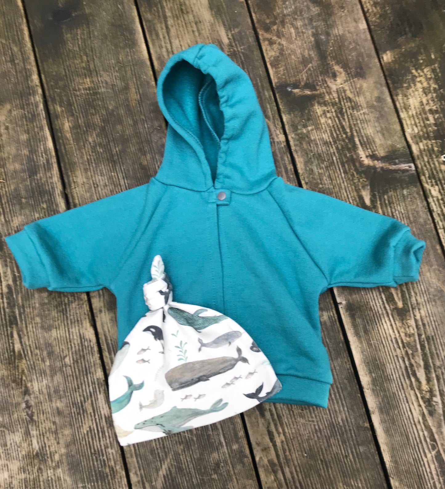 Teal hoodie and whale-patterned baby hat on a wooden surface