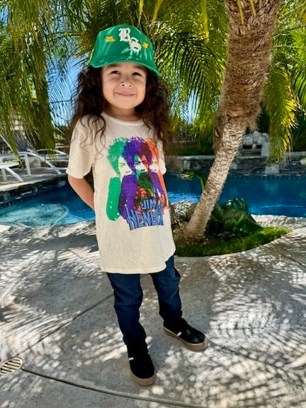 Child standing outside under a palm tree wearing a green baseball cap and a white Jimi Hendrix short sleeve tee