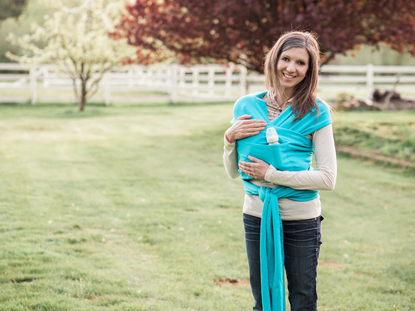 Woman holding a turquoise baby wrap in a grassy field with trees in the background