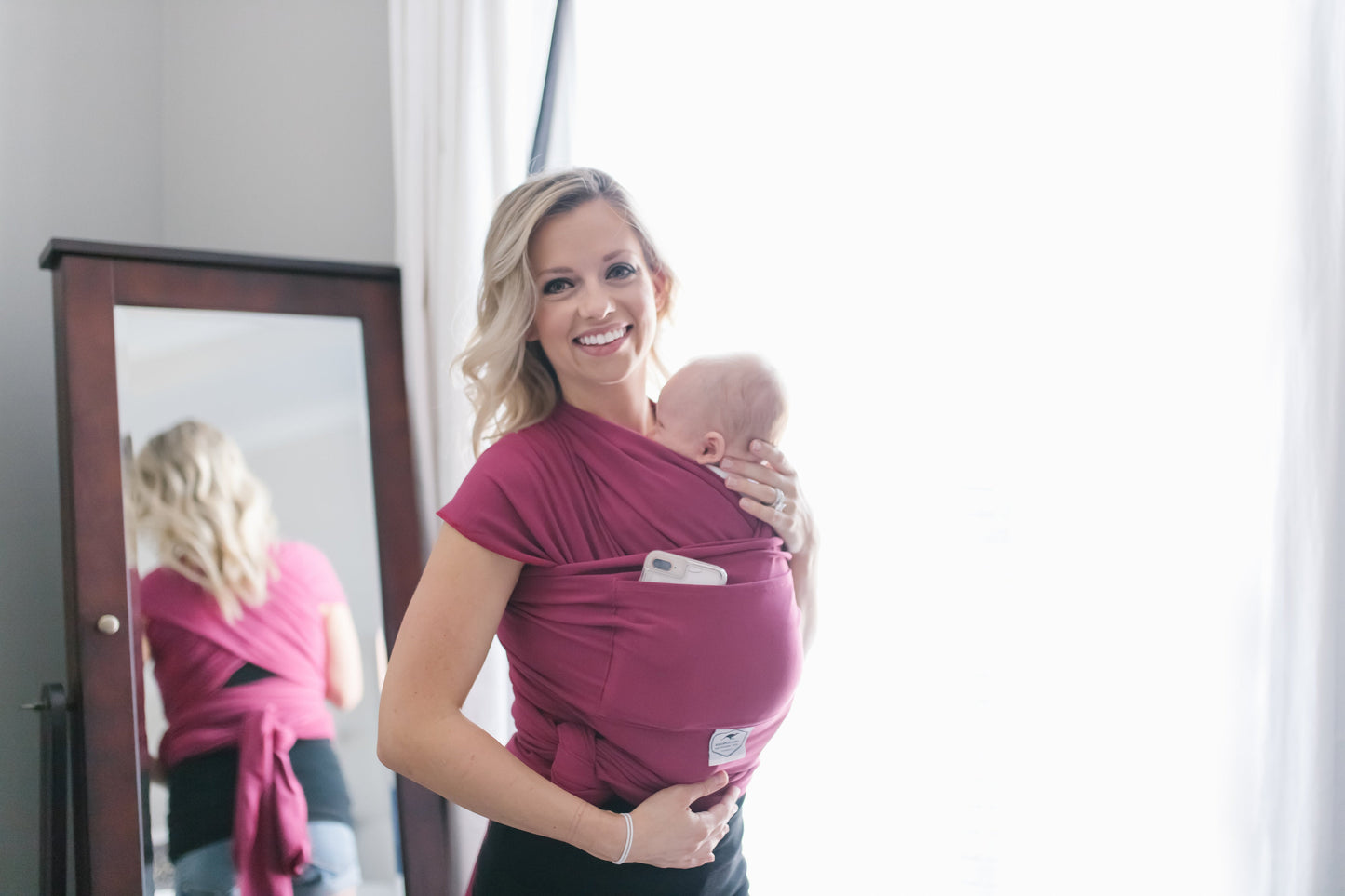 Woman holding a baby in a pink wrap in front of a mirror.
