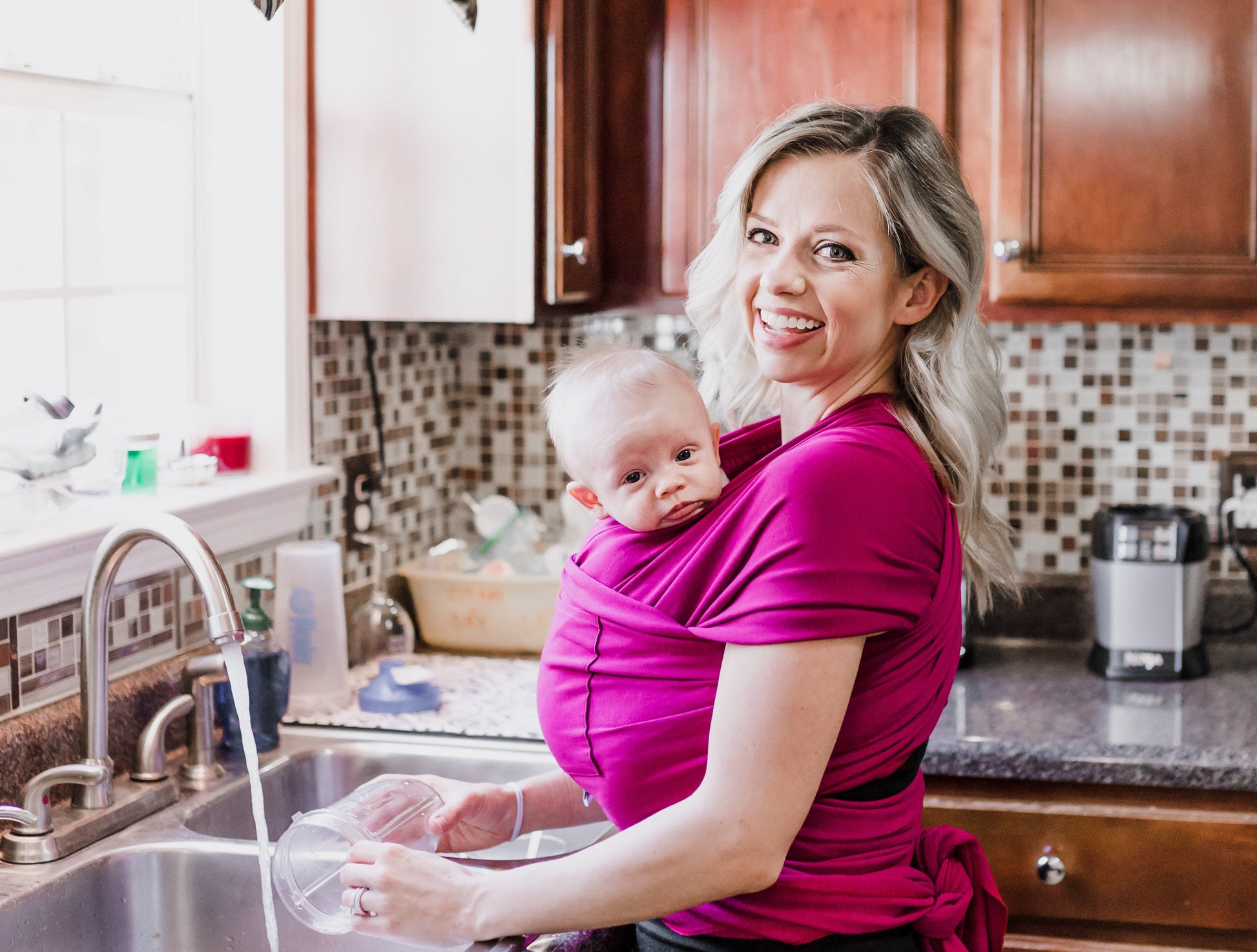 Woman in a kitchen holding a baby in a sling, washing dishes.