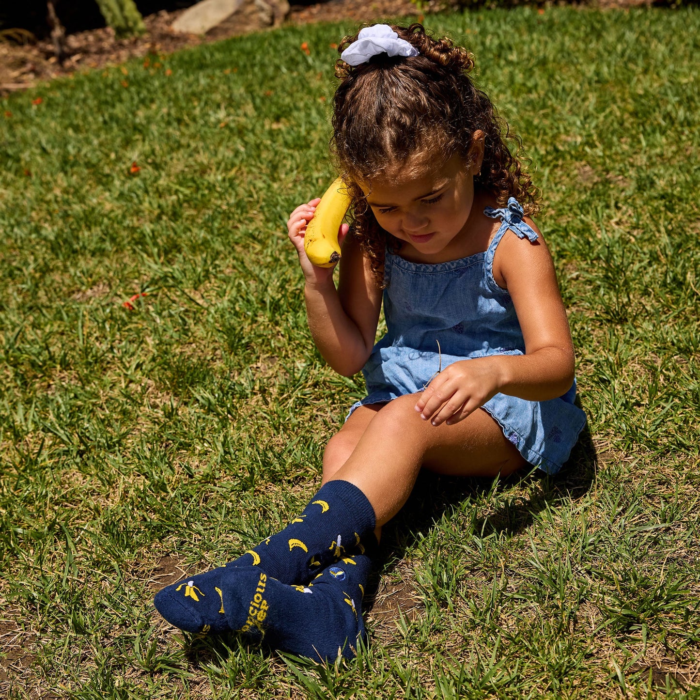 Young girl sitting on grass holding a banana, wearing blue dress and socks with yellow designs.