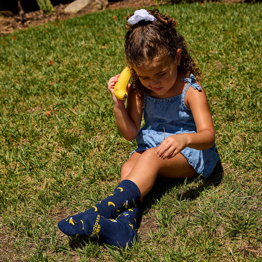 Young girl sitting on grass holding a banana, wearing blue dress and socks with yellow designs.