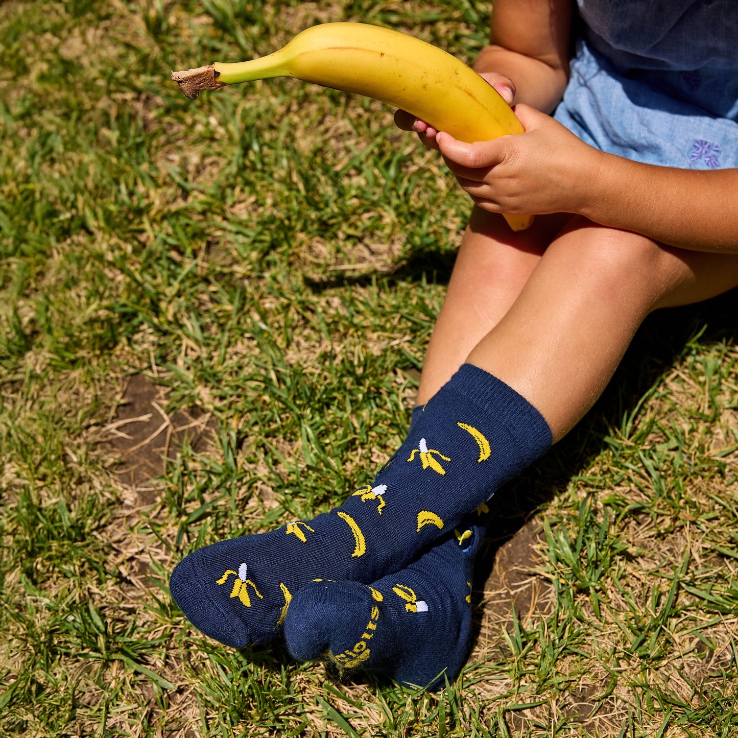 Person wearing navy blue socks with banana pattern sitting on grass holding a banana