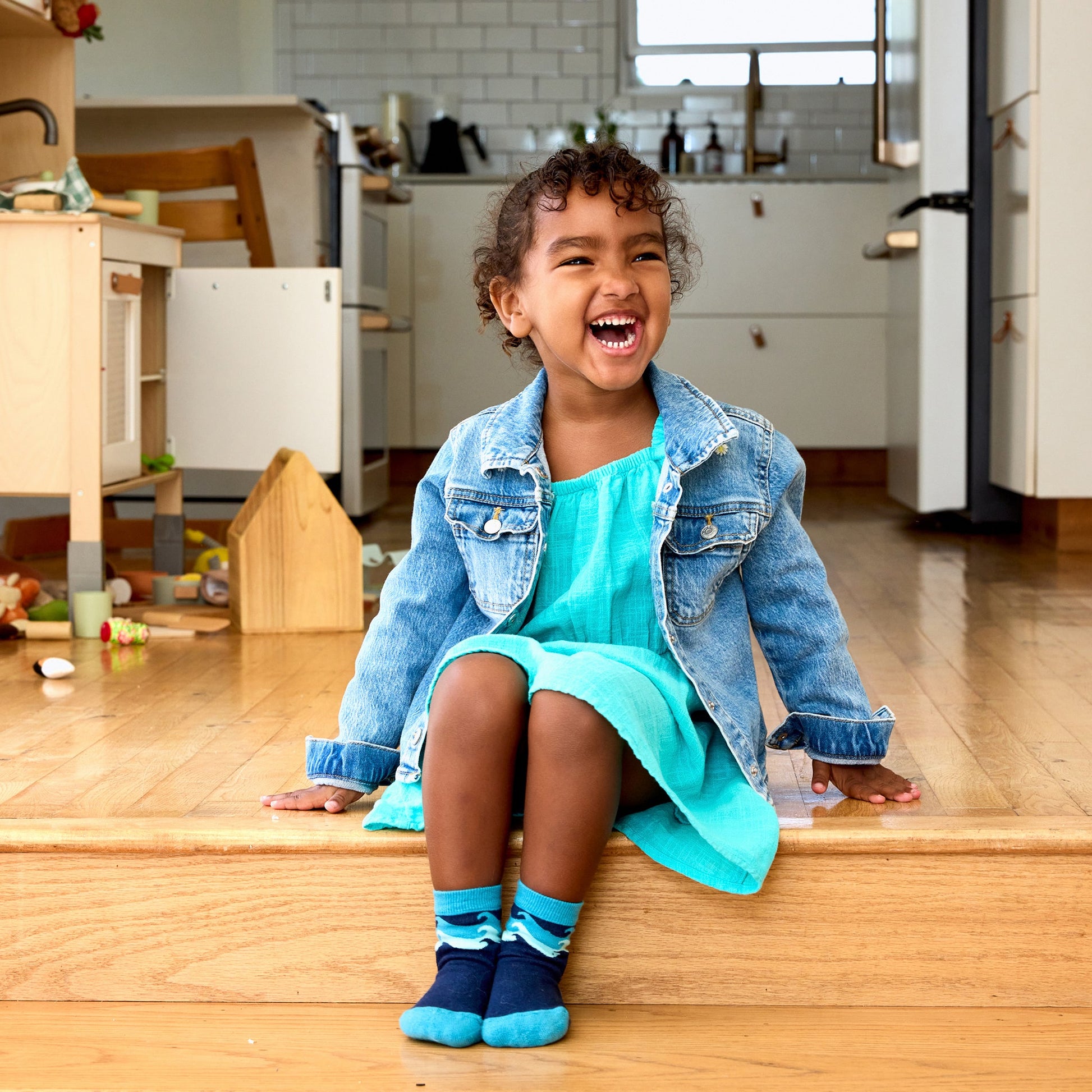 Child sitting on a kitchen counter wearing a blue dress and denim jacket, with a kitchen in the background.