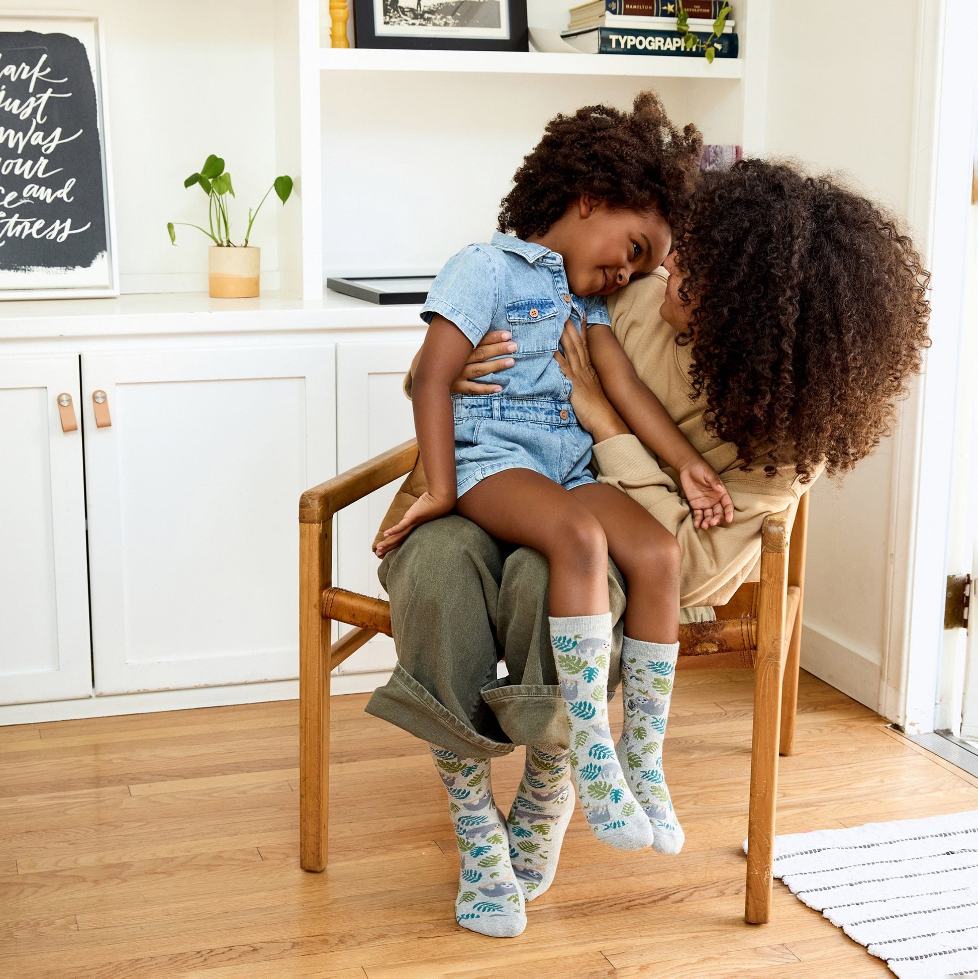 Two children sitting on a chair in a home setting with a shelf in the background.