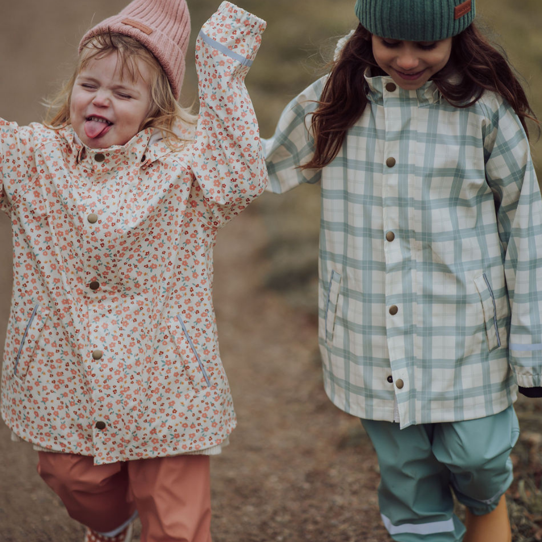 Two children in raincoats and hats standing outdoors on a grassy area.