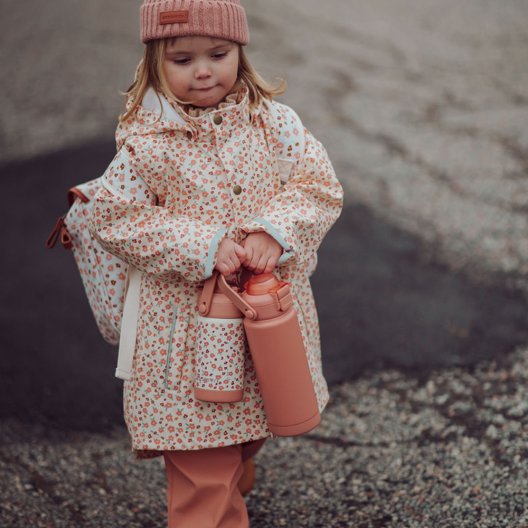 Child in a floral coat holding a pink thermos outdoors