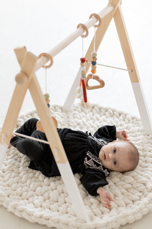 Baby lying on a white rug with a wooden play gym and hanging toys.