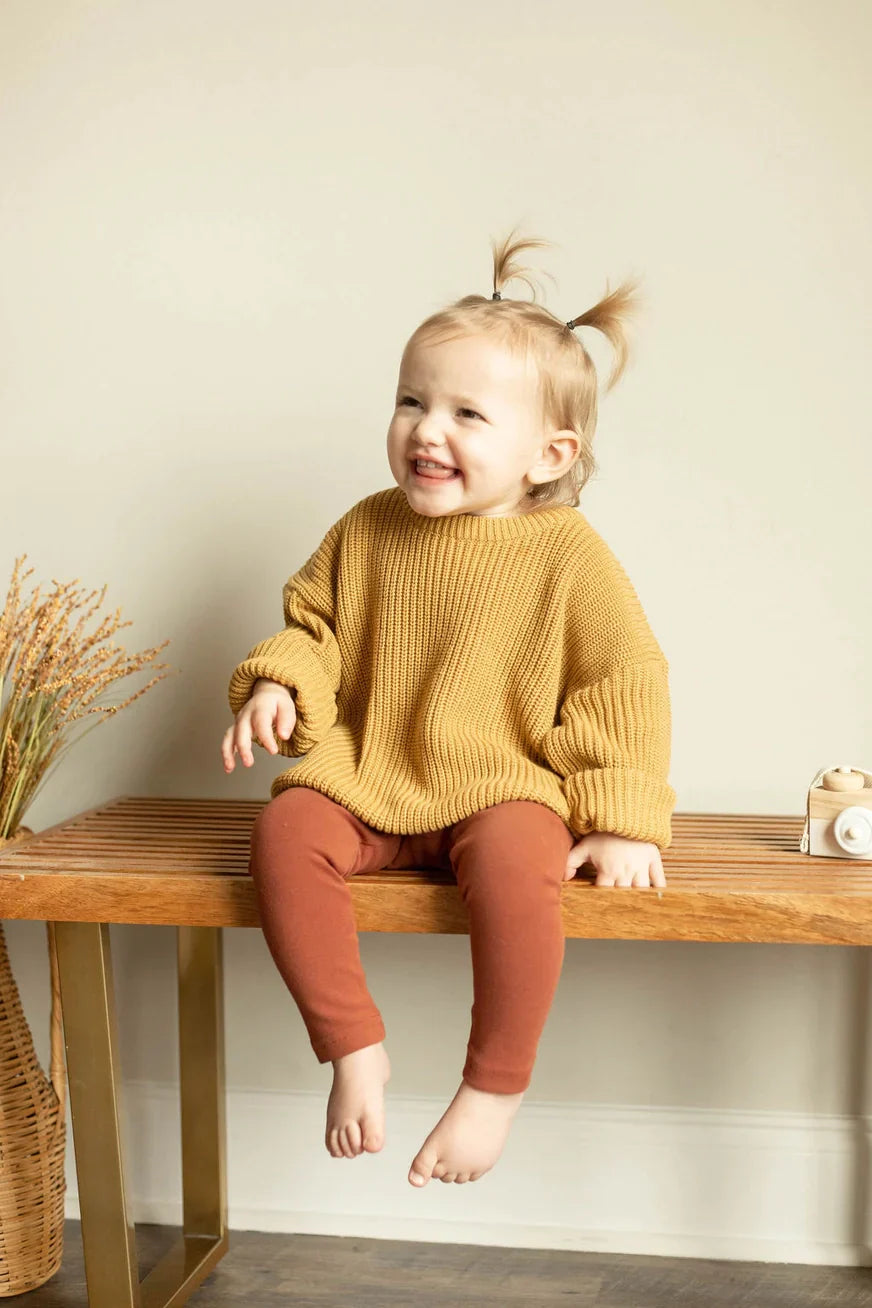 Child sitting on a wooden bench wearing a mustard yellow sweater and brown pants.