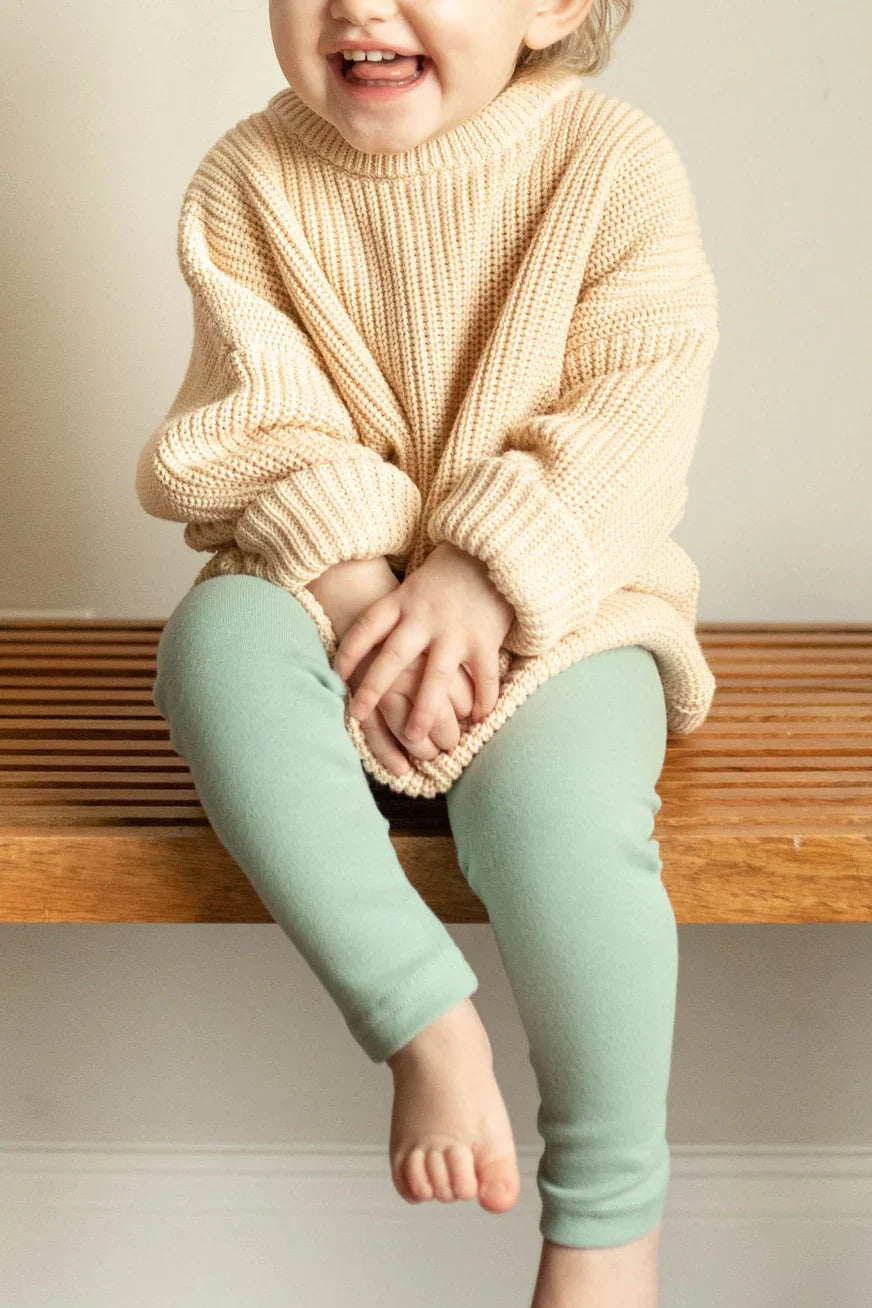 Child wearing a beige sweater and light green leggings sitting on a wooden bench.
