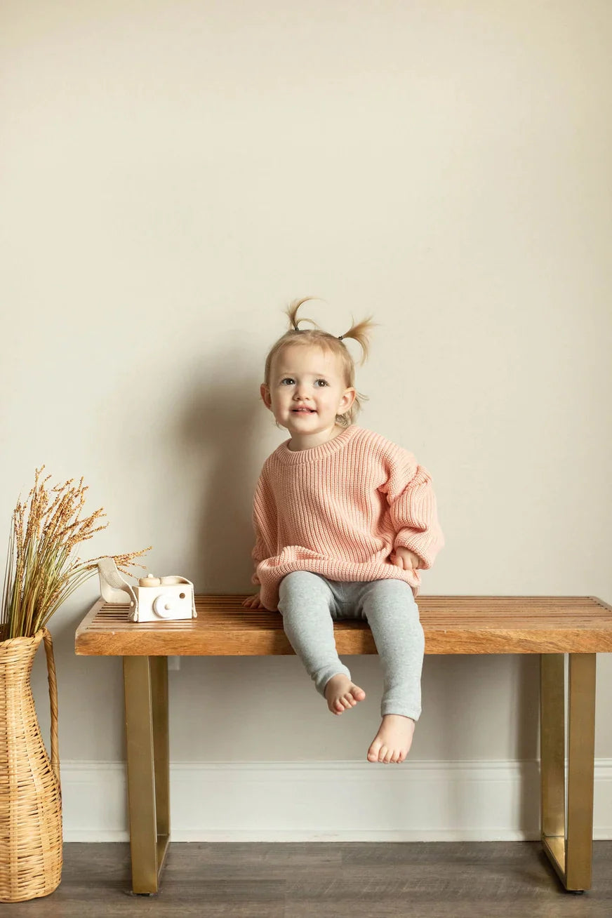 Child sitting on a wooden bench against a plain wall with a decorative plant to the side.