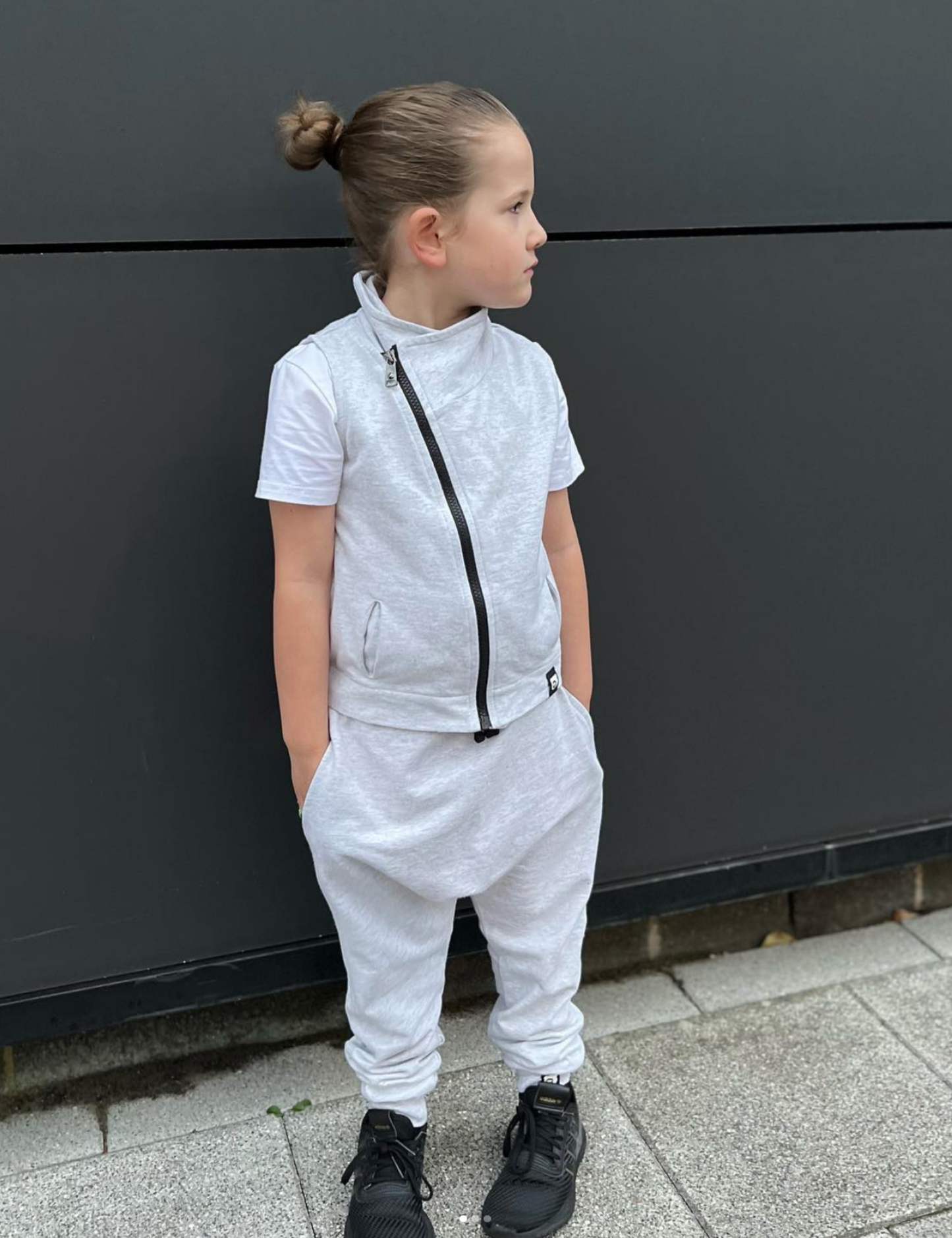 Child wearing a gray tracksuit standing against a dark wall.