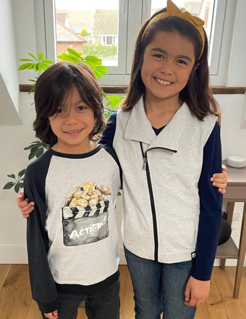 Two children standing together indoors, smiling at the camera.