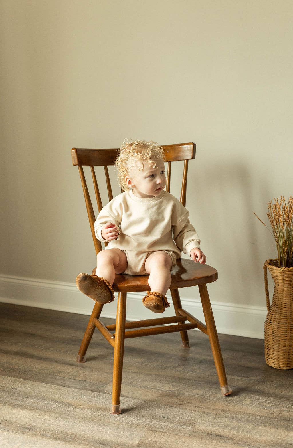 Child sitting on a wooden chair in a room with a neutral wall and a basket of dried plants.