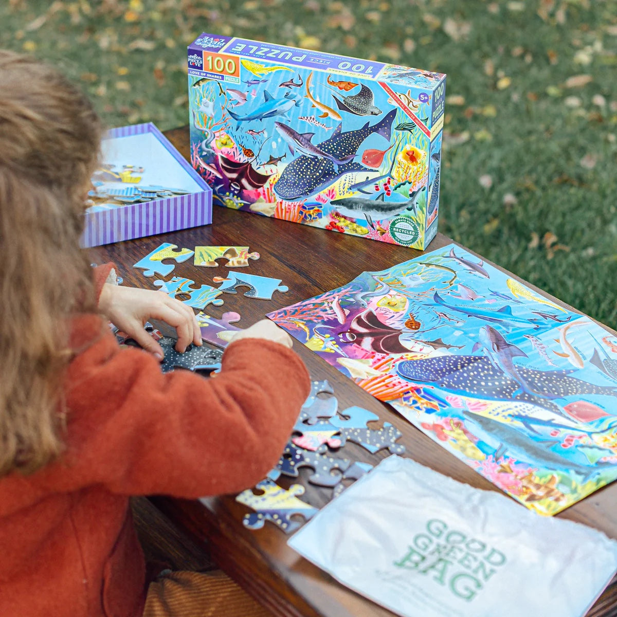 Child wearing an orange sweater making 100-piece Love of Sharks puzzle on a wooden table outside