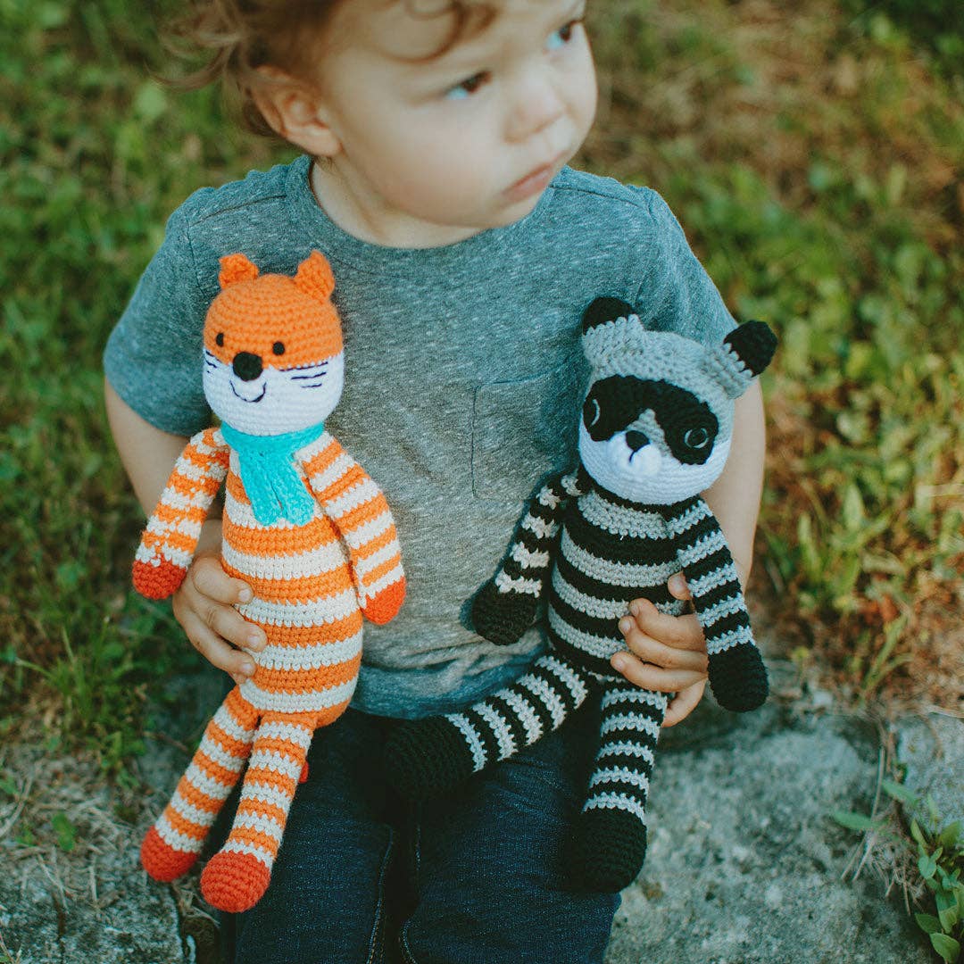 Child holding two crocheted animals, one orange and white striped fox and one black and white striped raccoon, outdoors.