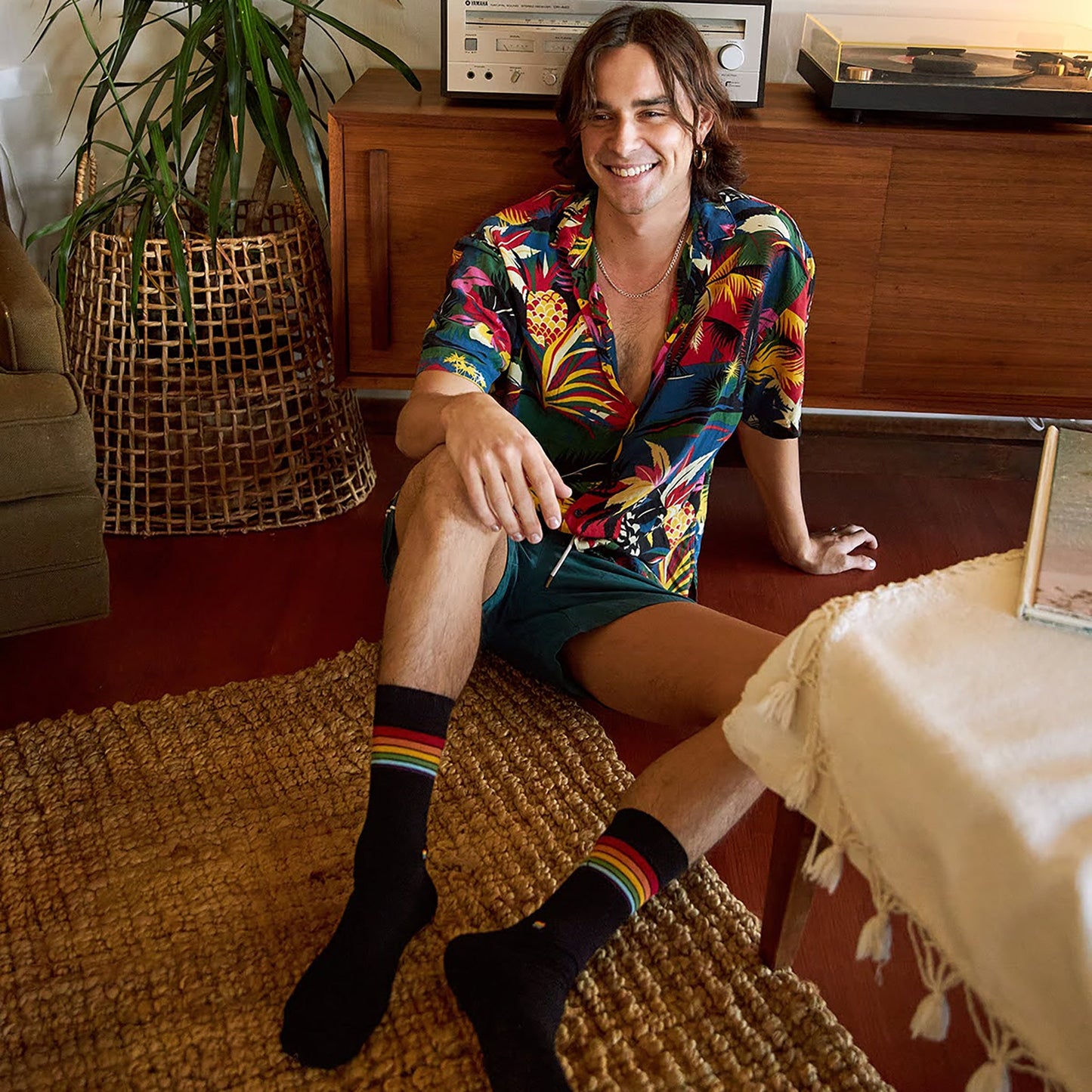 Man sitting on a rug in a living room wearing a colorful shirt and rainbow socks.