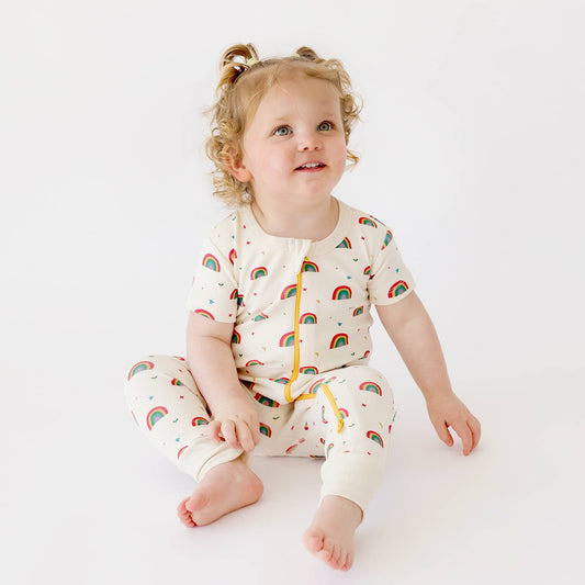 Toddler with curly blonde hair with two pig tales sitting and looking up while wearing parade organics Short Sleeve Rainbow Romper w/ 2-way Zipper