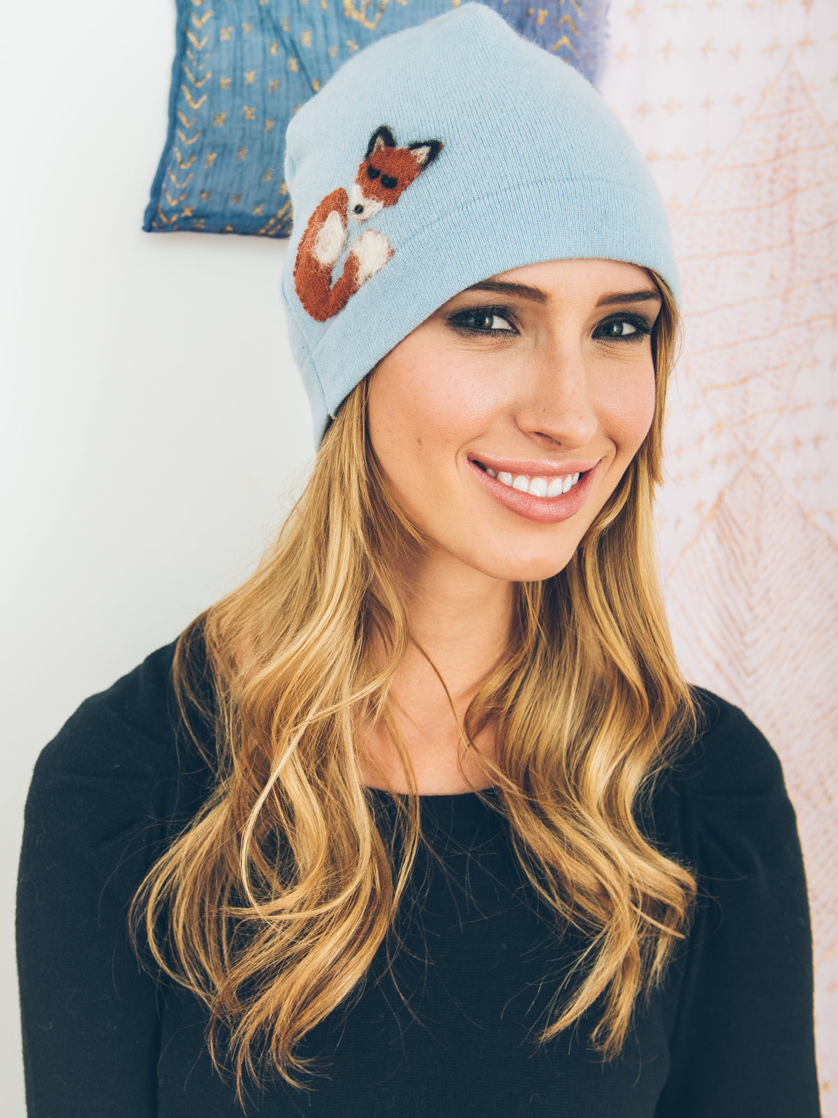 Woman wearing a light blue beanie with a fox design, smiling against a neutral background.