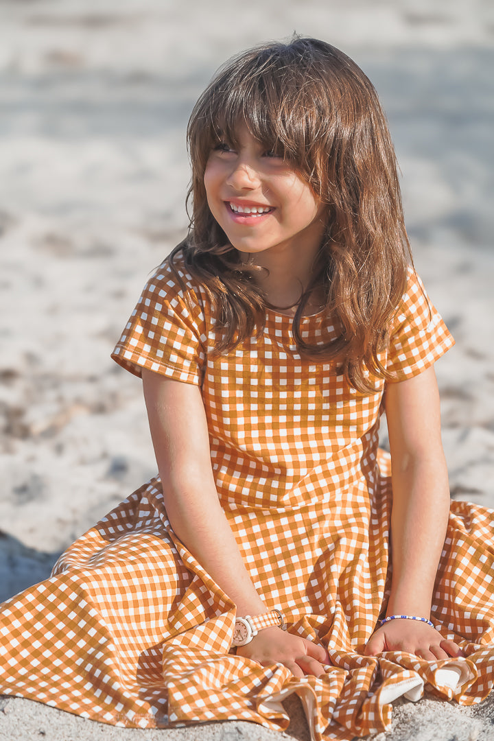 Young girl in a yellow checkered dress sitting on a sandy beach.