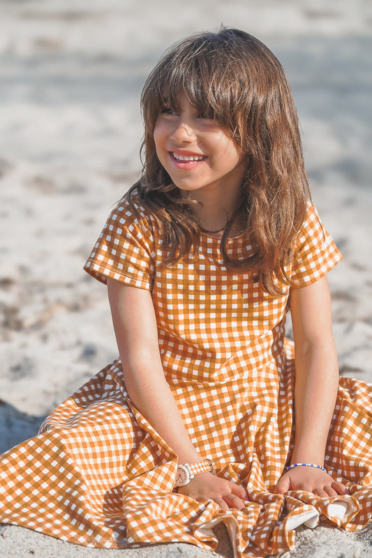 Young girl in a yellow checkered dress sitting on a sandy beach.