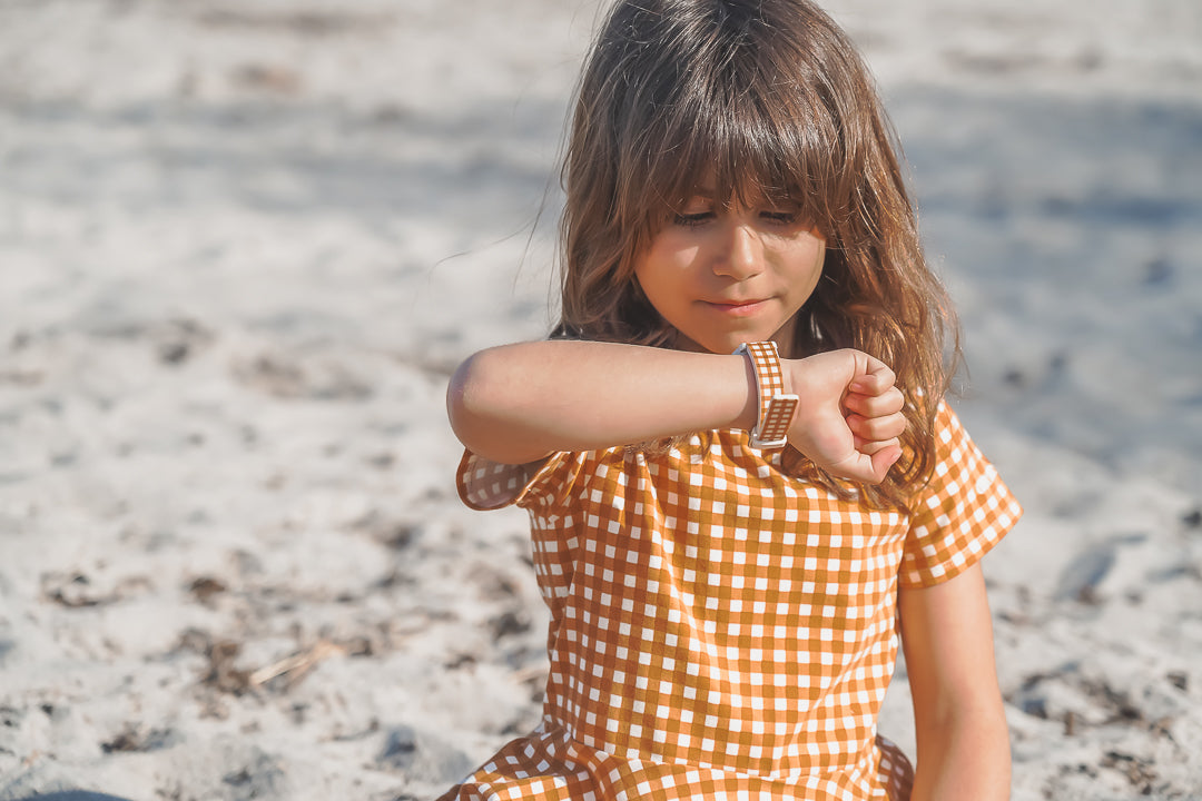 Child in a checkered dress sitting on a sandy beach.