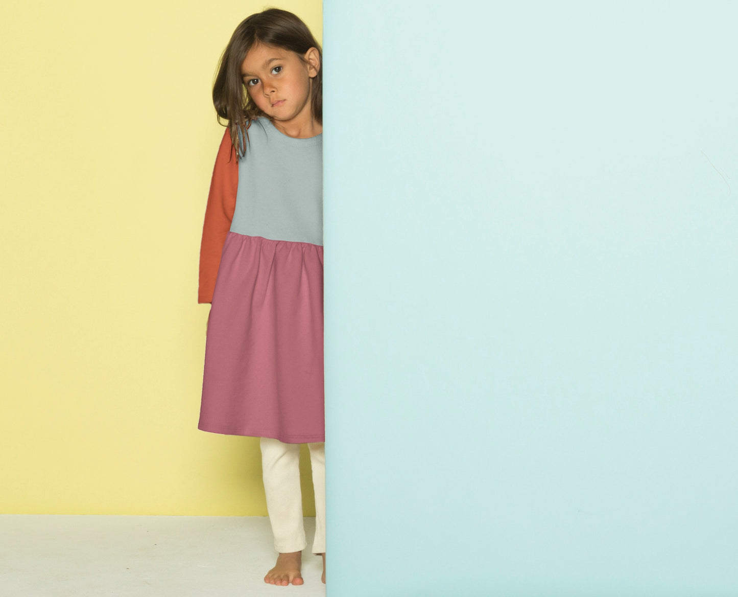 Young girl standing between a yellow and blue background wearing a two-tone dress.