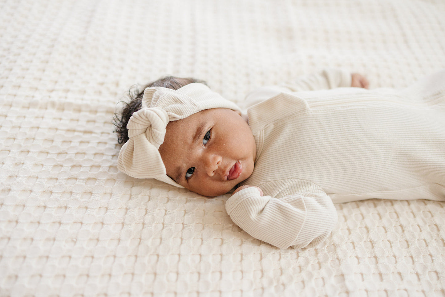 Newborn baby wrapped in white with a headband, lying on a textured white blanket.