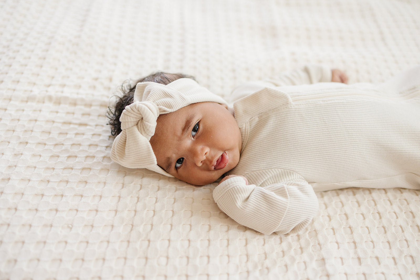Newborn baby wrapped in white with a headband, lying on a textured white blanket.