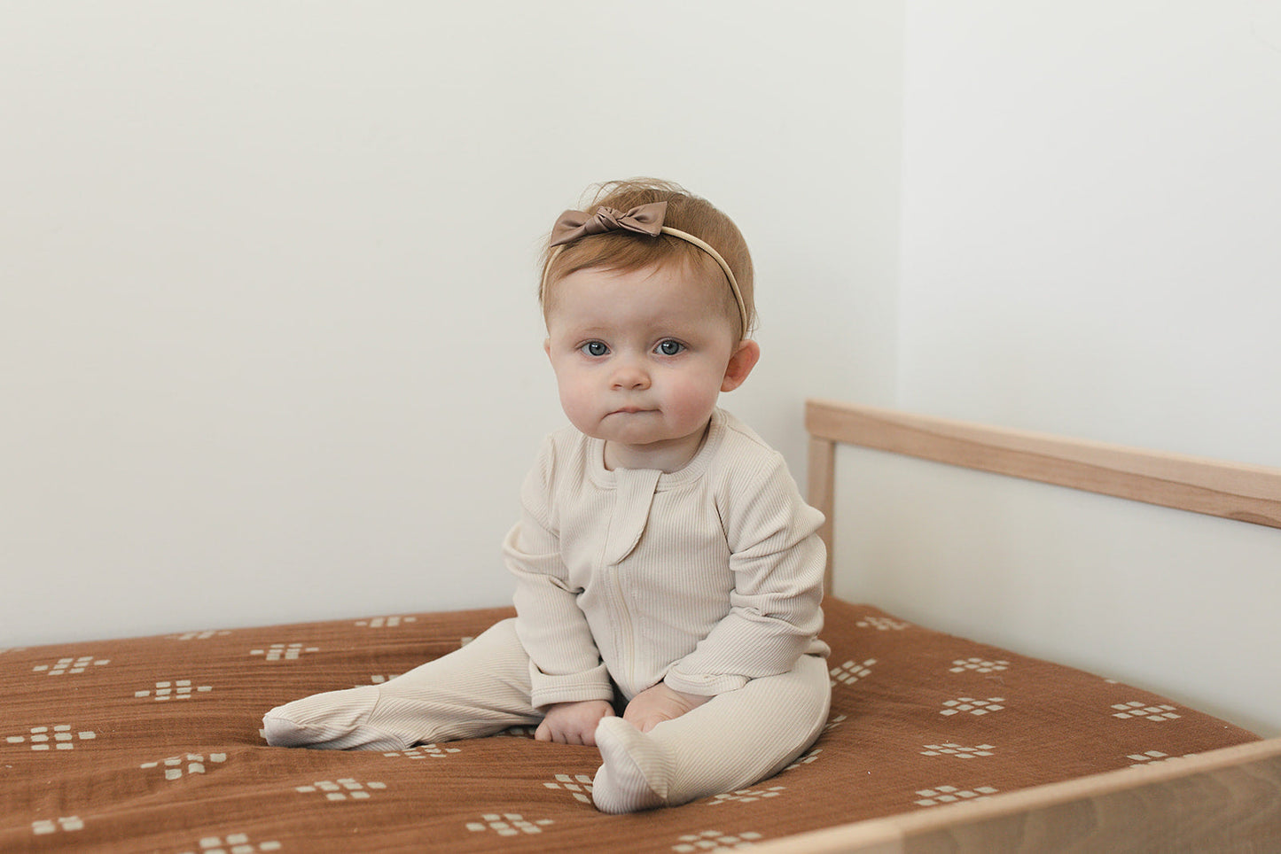 Baby sitting on a bed wearing a beige outfit and headband.