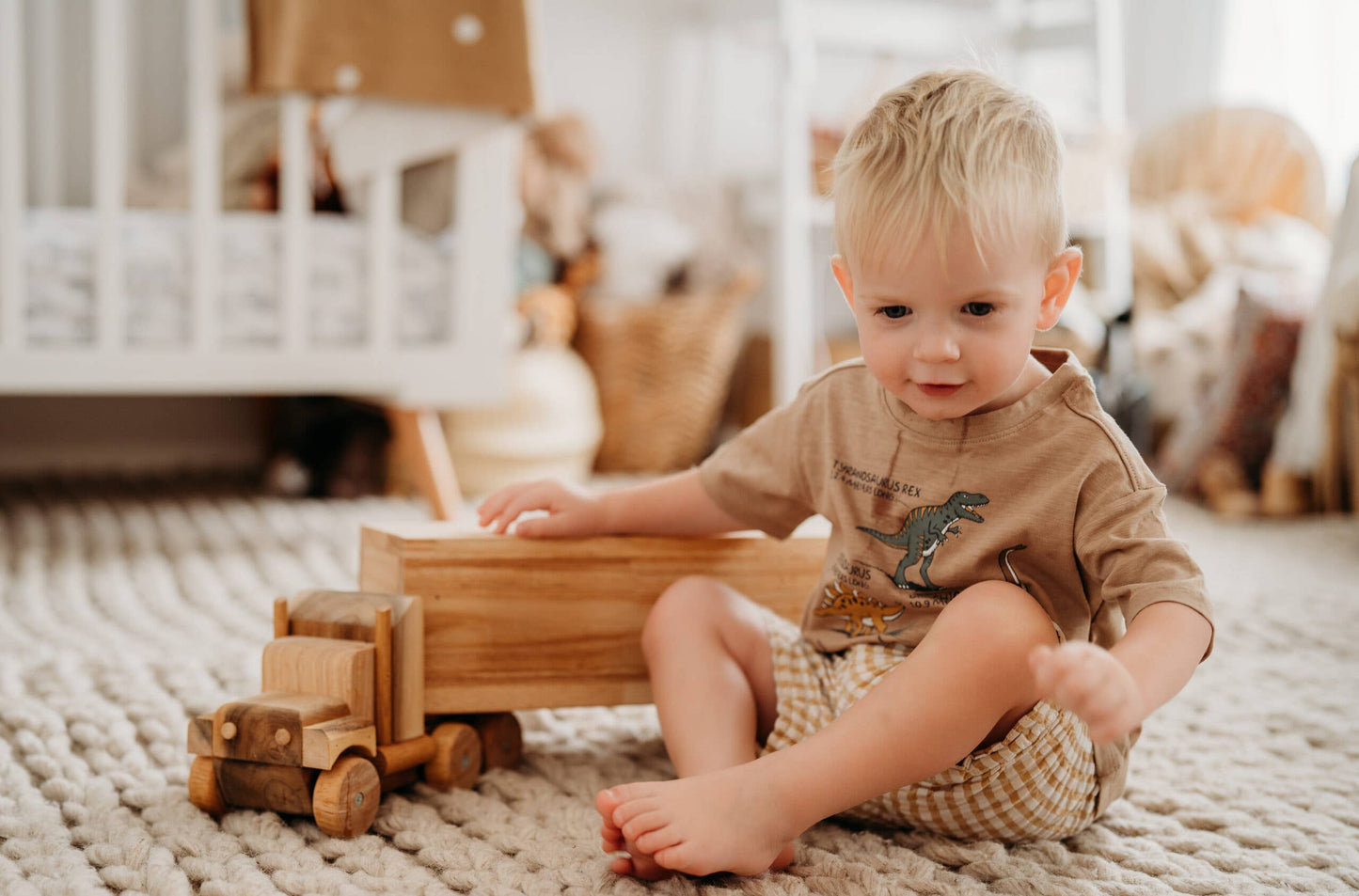Blonde short haired child in a tan shirt sitting on a light colored rug in a child's room playing with a wooden cargo truck