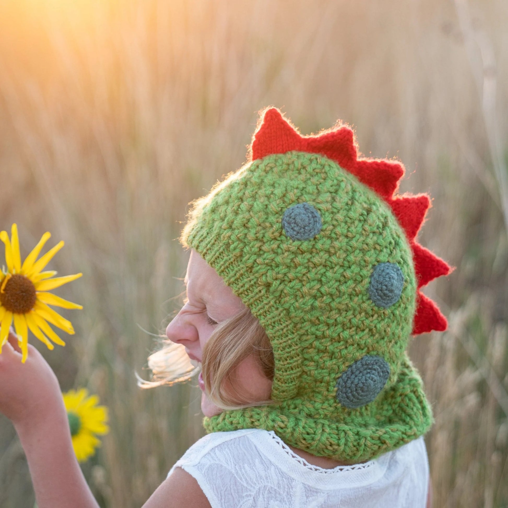 Child smiling holding a sunflower while sitting in a field and wearing wool dinosaur hood