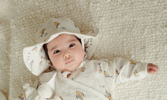 A baby with fair skin brown hair and brown eyes laying on a white textured blanket.  the baby is wearing a matching sunhat and crossover romper that are white and have yellow and blue kites printed all over..