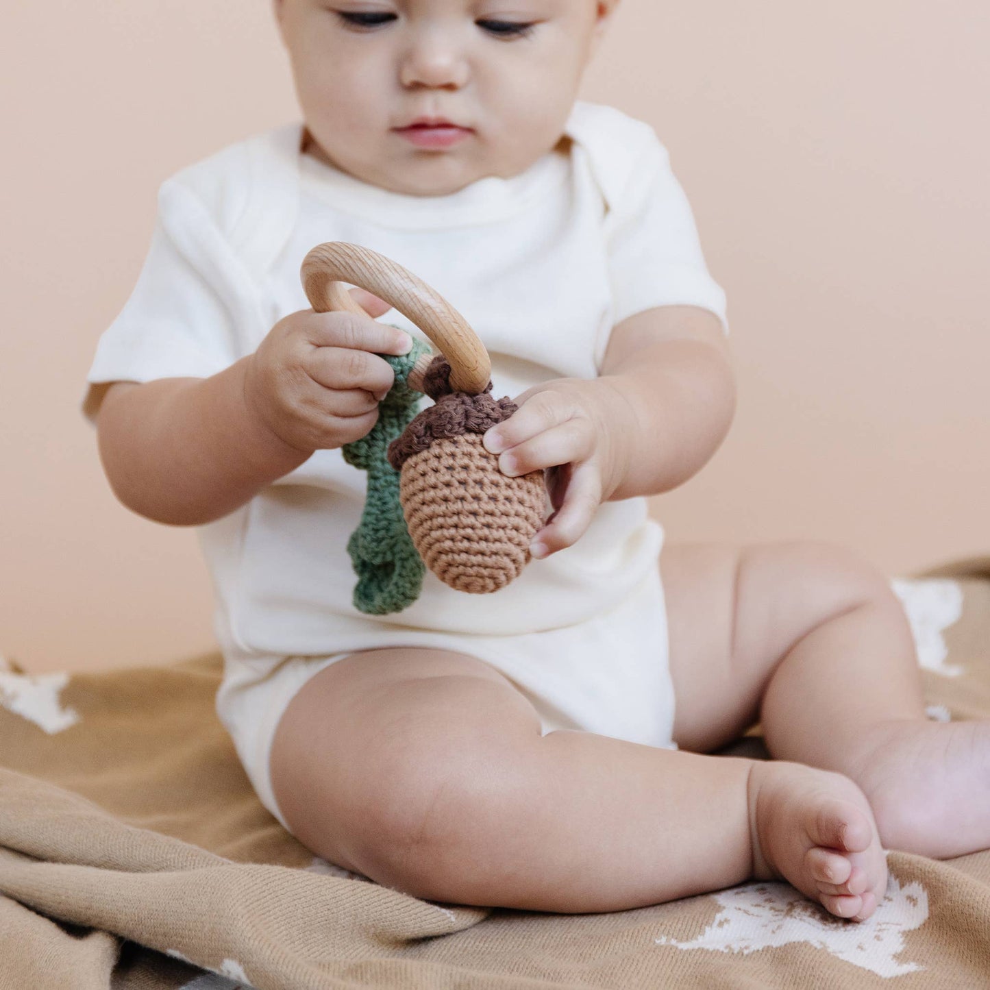 Baby sitting on a beige blanket holding a wooden teether with knitted acorn and leaf