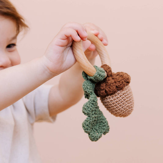 Child holding a wooden teething ring with knitted acorn and leaf against a soft pink background