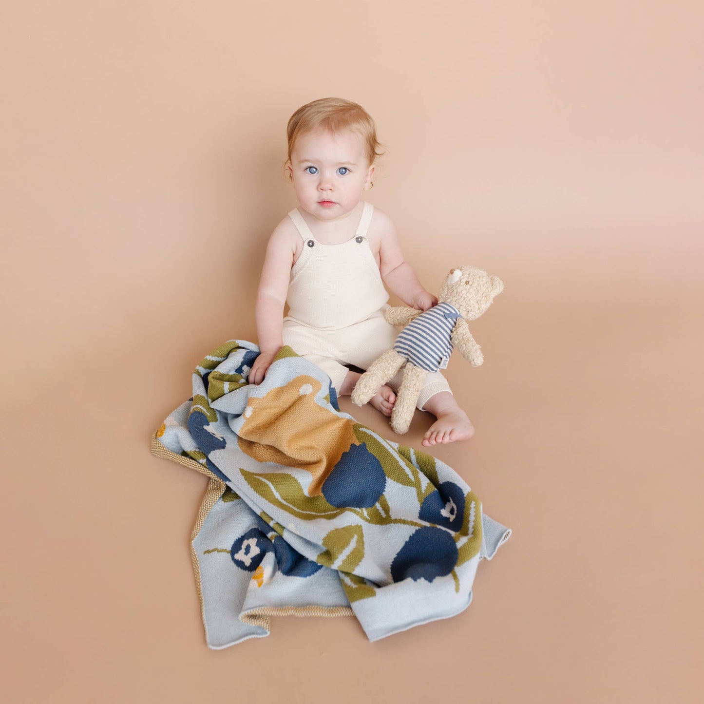 Baby sitting on a blueberry blanket holding a bear toy, with a beige background