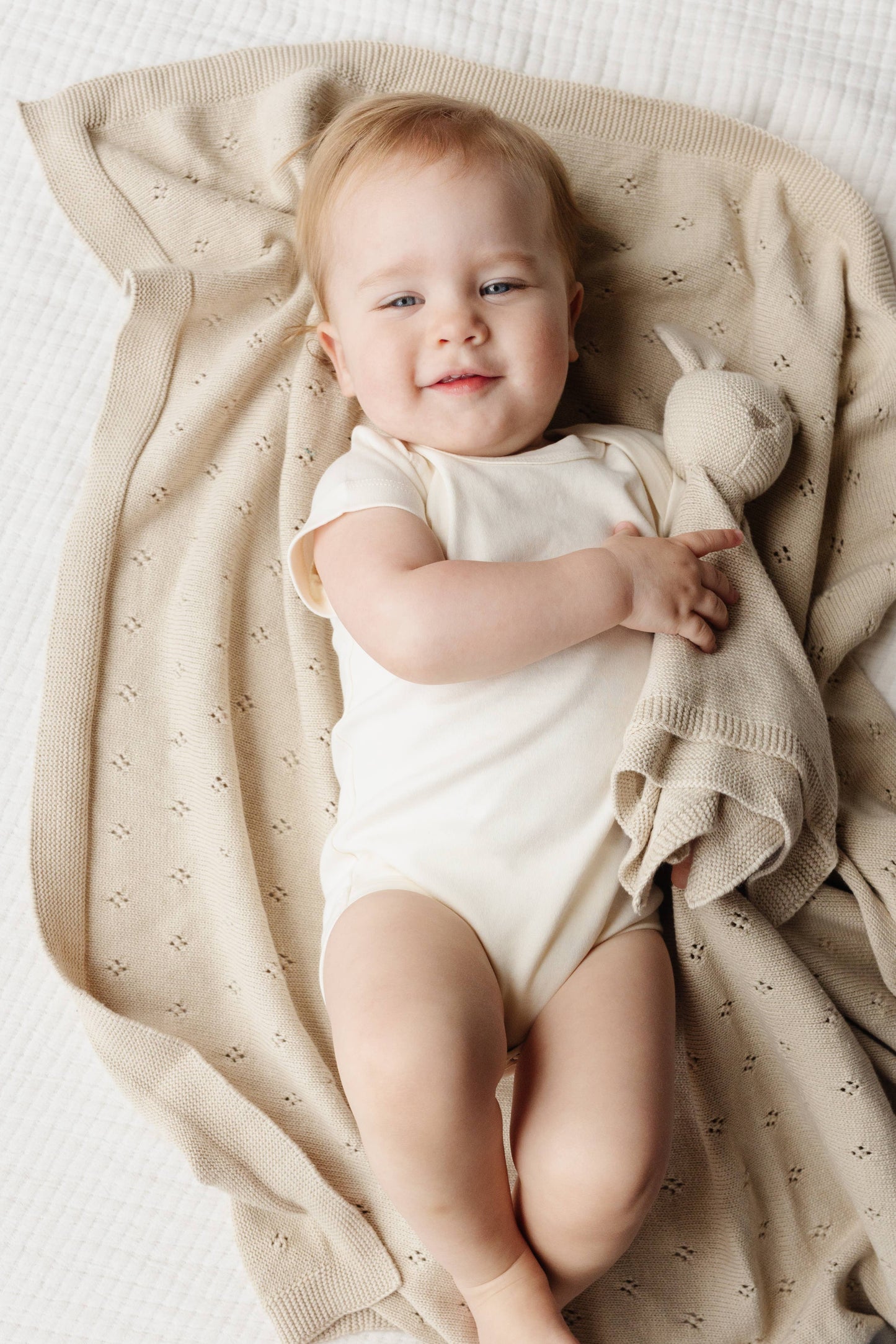 Baby in a white onesie lying on a beige blanket with a bunny toy