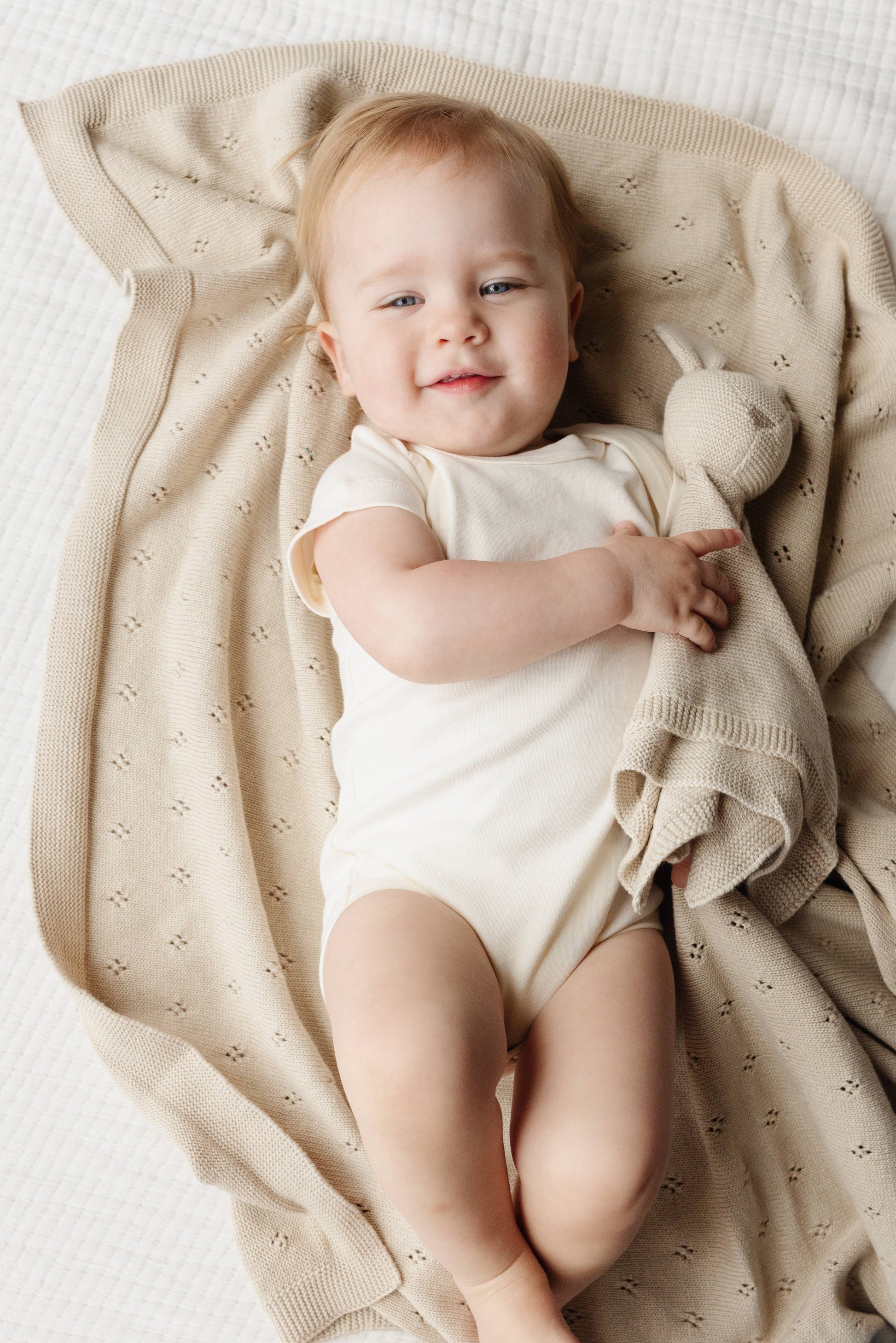 Baby in a white onesie lying on a beige blanket with a bunny toy