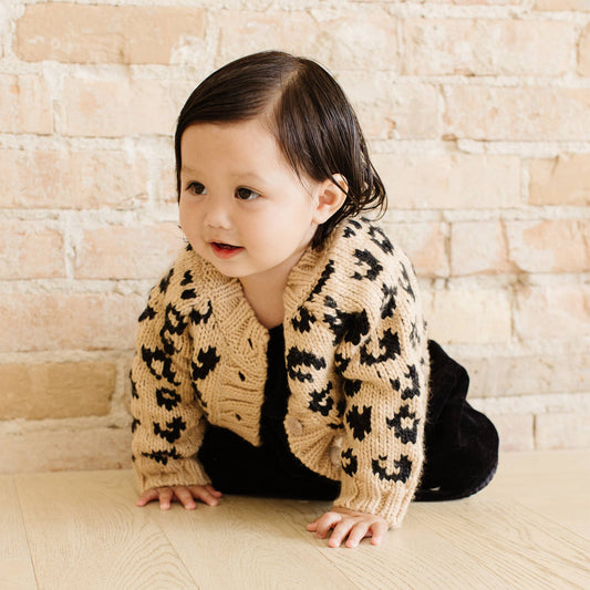 Child wearing a leopard print cardigan sitting on the floor against a brick wall.