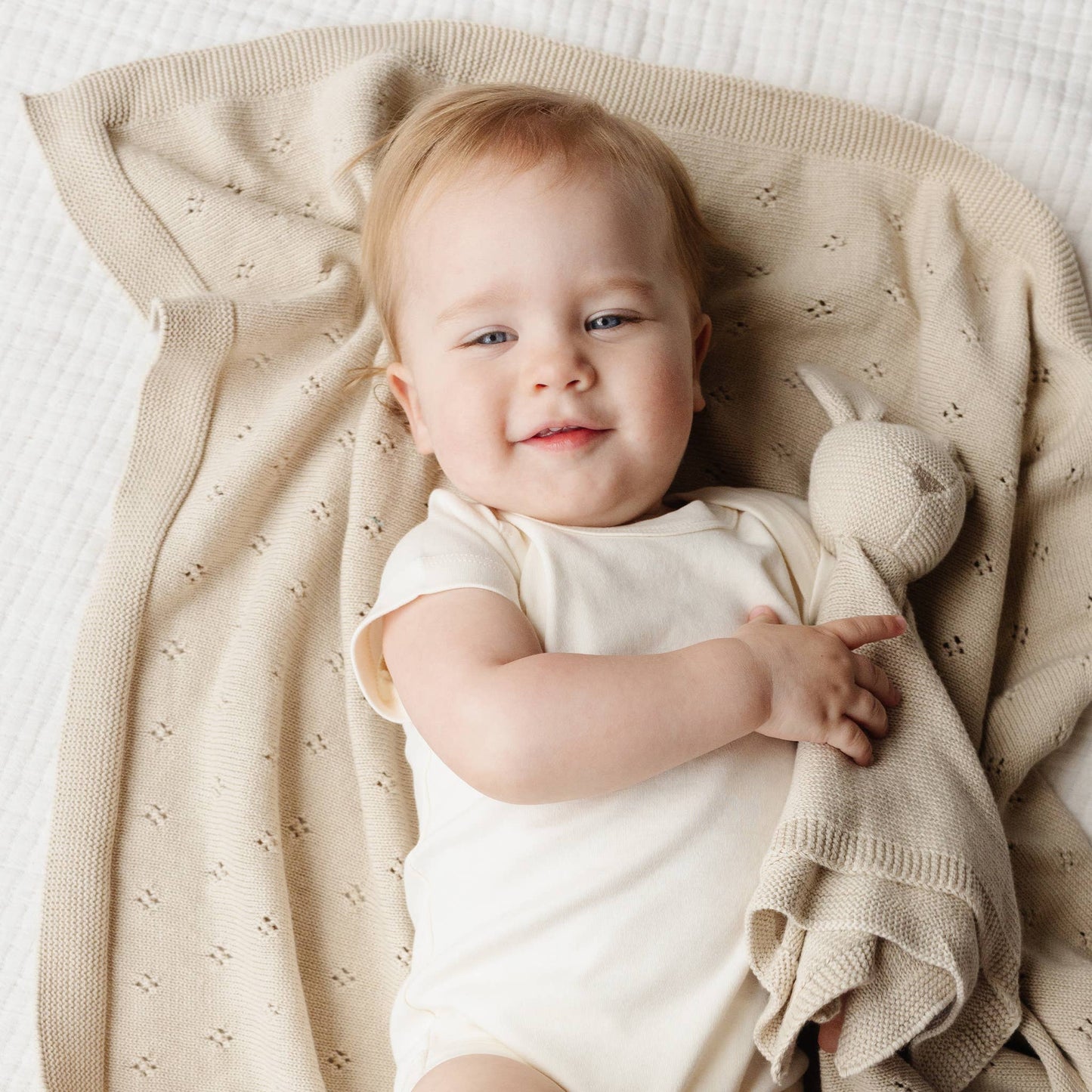 Baby lying on a beige blanket with a bunny toy, wearing a white onesie.