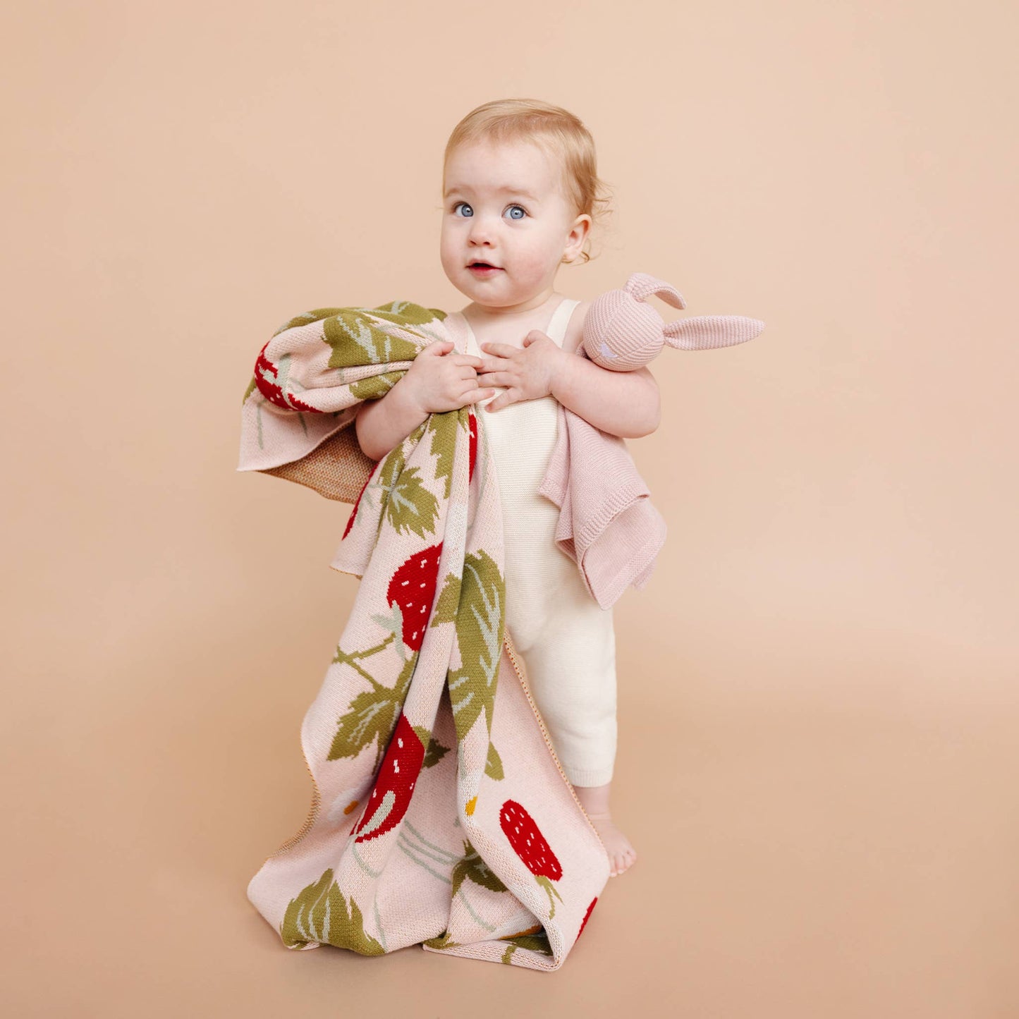 Baby holding a strawberry-patterned blanket and a pink toy against a beige background