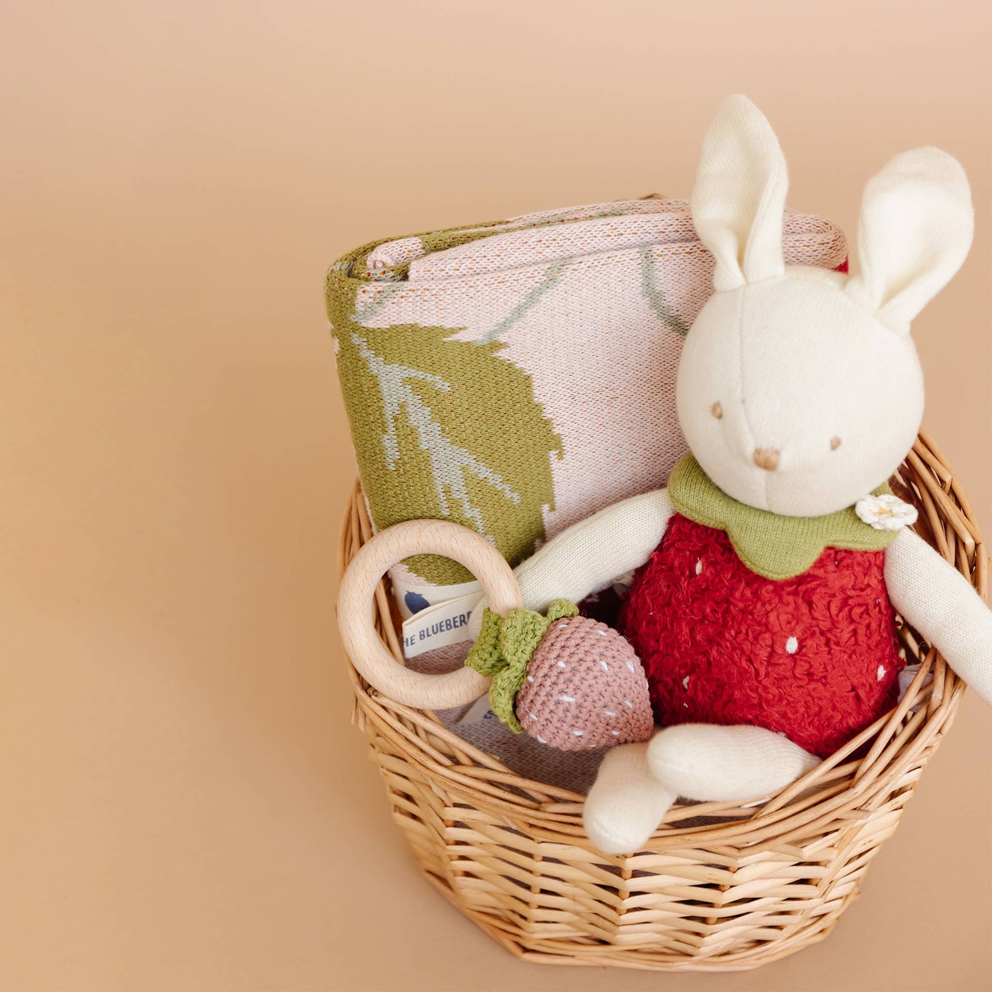 Wicker basket with a plush strawberry rabbit toy, strawberry blanket and wooden teether on a beige background